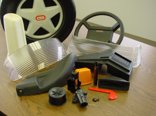 Collection of toy car parts including a wheel, steering wheel, dashboard, plastic body, and various small accessories on a wooden surface.