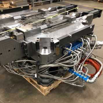 Large industrial metal mold or machine component with connected hydraulic or pneumatic hoses, placed on a wooden pallet in a factory setting.