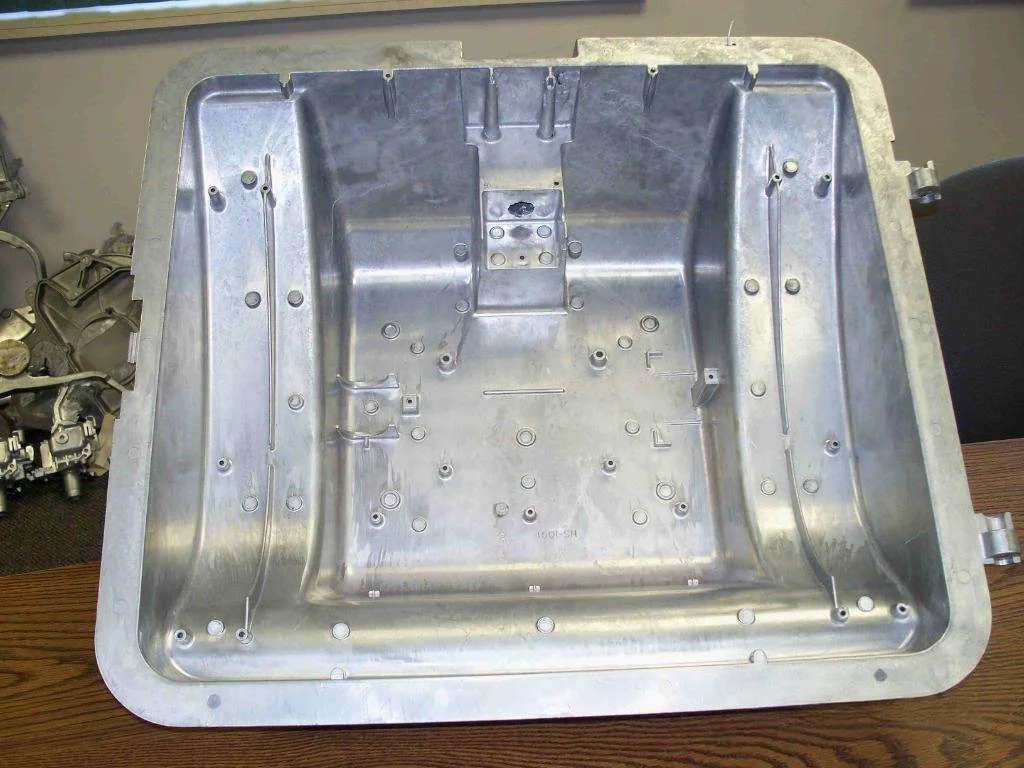 A metal mechanical or electronic enclosure or housing with multiple mounting points and holes on a wooden table.