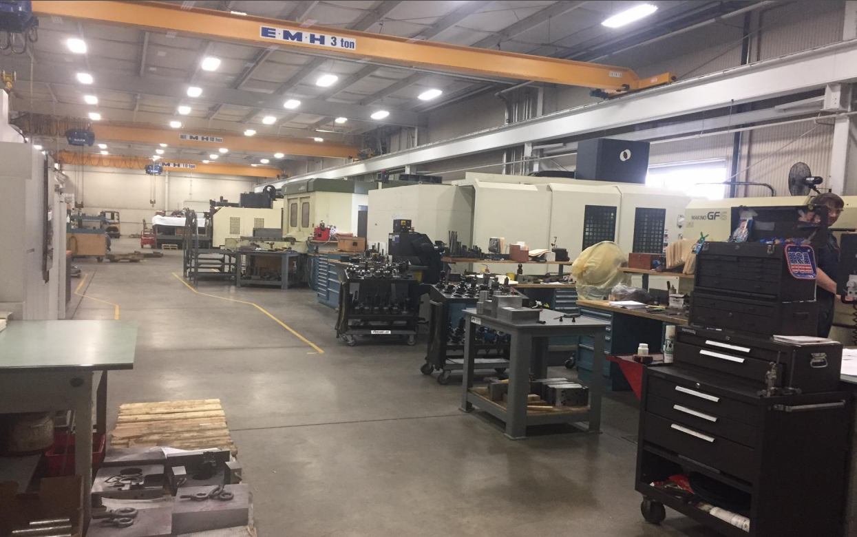 An industrial manufacturing or machine shop with various workbenches and machines, including a large CNC machine, tool carts, and overhead cranes, in a spacious warehouse.