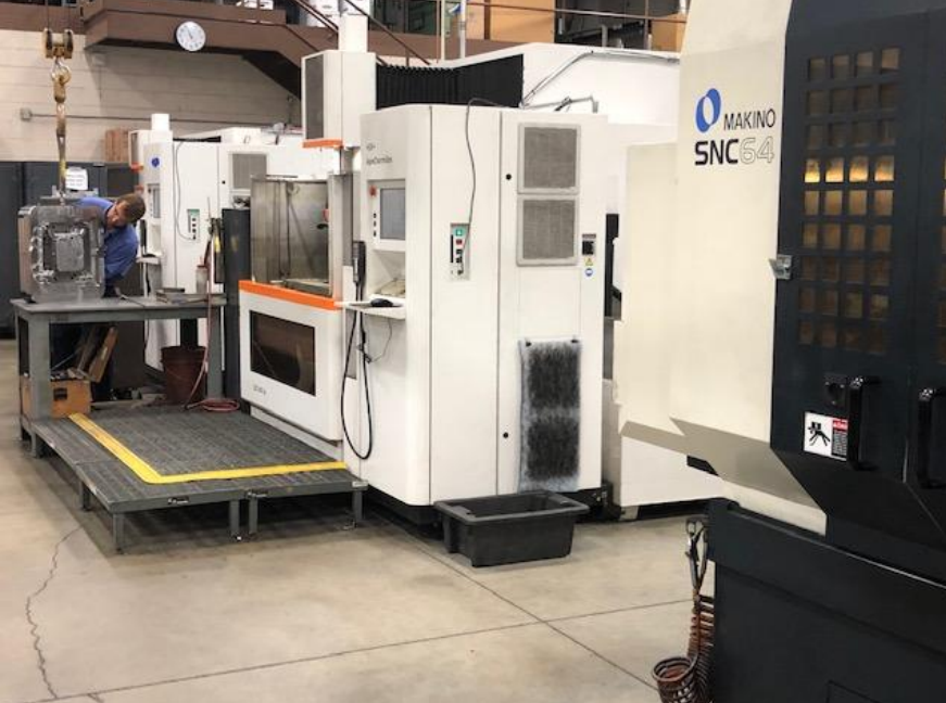 Industrial manufacturing setup with a large CNC machine labeled 'Makino SNC 64' and a worker in a blue shirt working on a smaller component on a nearby table.