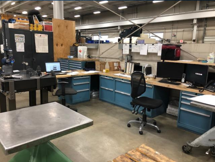 Industrial workspace with blue storage cabinets, office chairs, desktop computers, and boxes in a warehouse setting.
