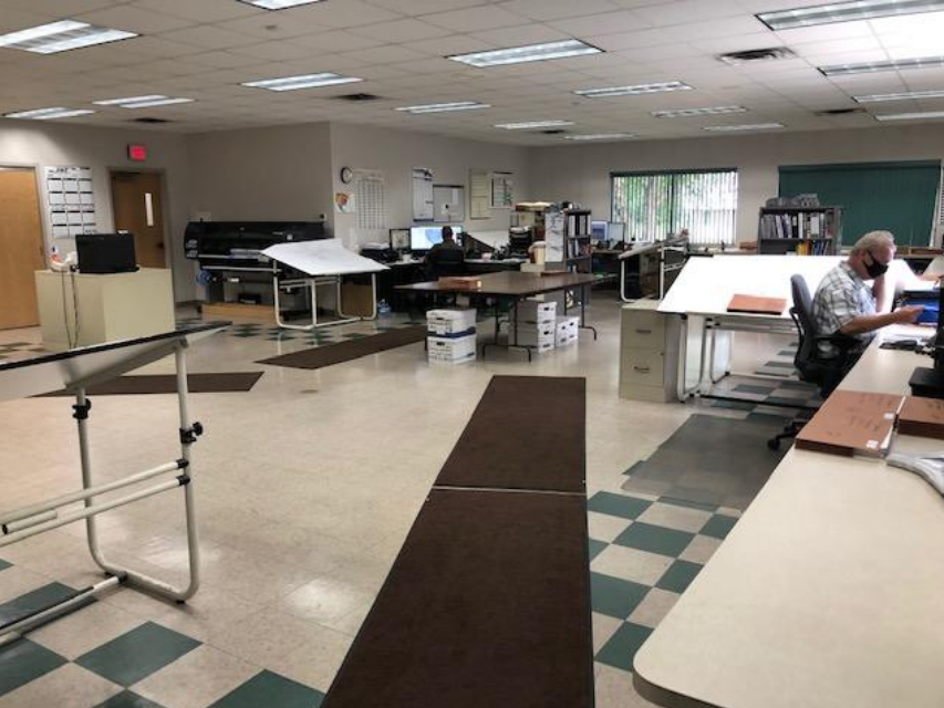 Office room with multiple desks, computers, and employees working, some wearing masks, with filing cabinets and windows in the background.