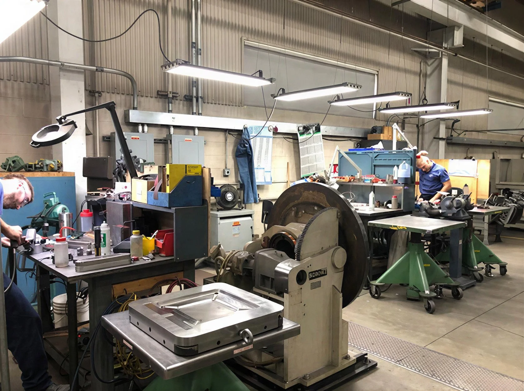 An industrial workshop with various machines and tools on workbenches, illuminated by overhead lights. Two workers are present, one on the left working with equipment, and one on the right wearing a blue shirt and a red cap, working near a large circ