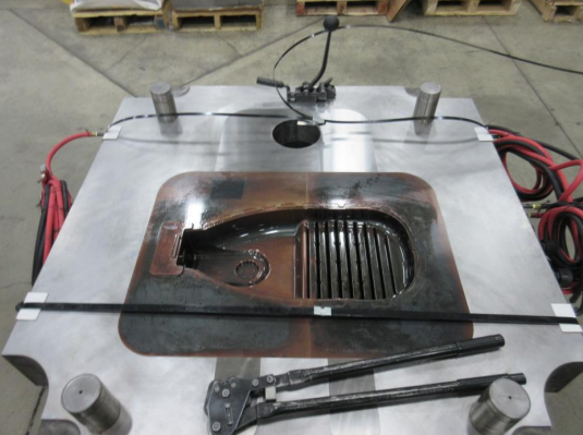 A stainless steel welding table with a cutout in the center, surrounded by welding equipment, clamps, and cables in an industrial workspace.