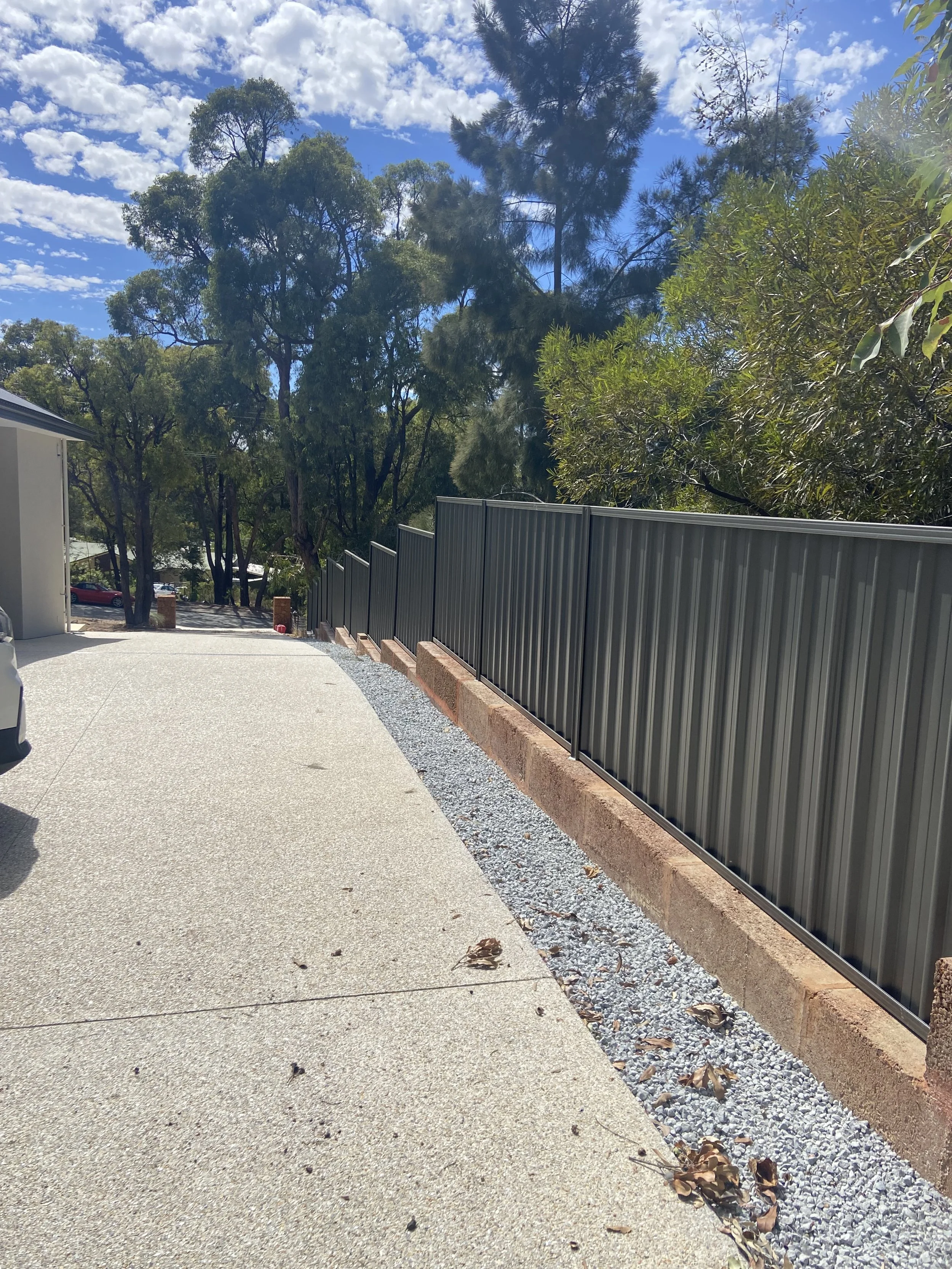 A driveway with a gravel border and a black metal fence, flanked by trees and a cloudy blue sky.