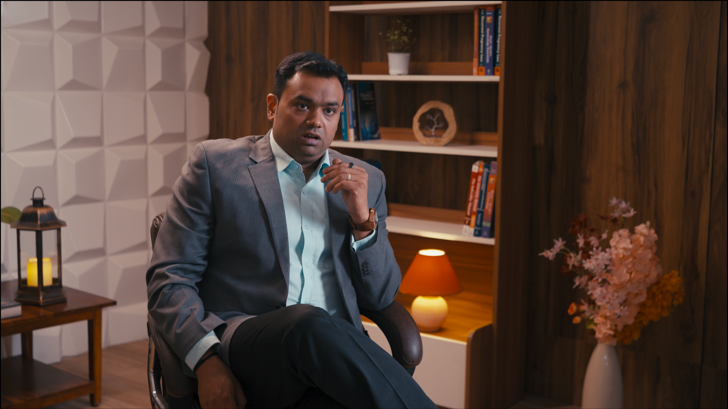 A man in a suit sitting in a modern office with decorative shelves, a small potted plant, a wooden sculpture, and pink flowers in a vase.