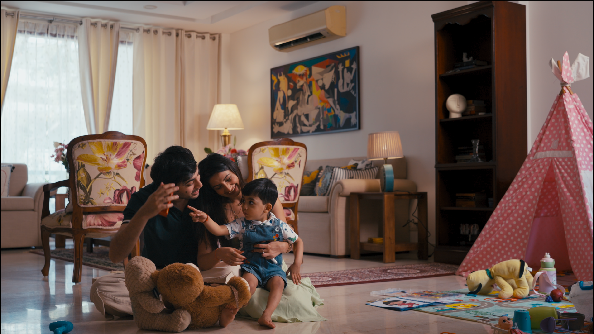 A family of three, including a man, woman, and young boy, playing and laughing together on the floor of a living room filled with toys, chairs, and a teepee tent.