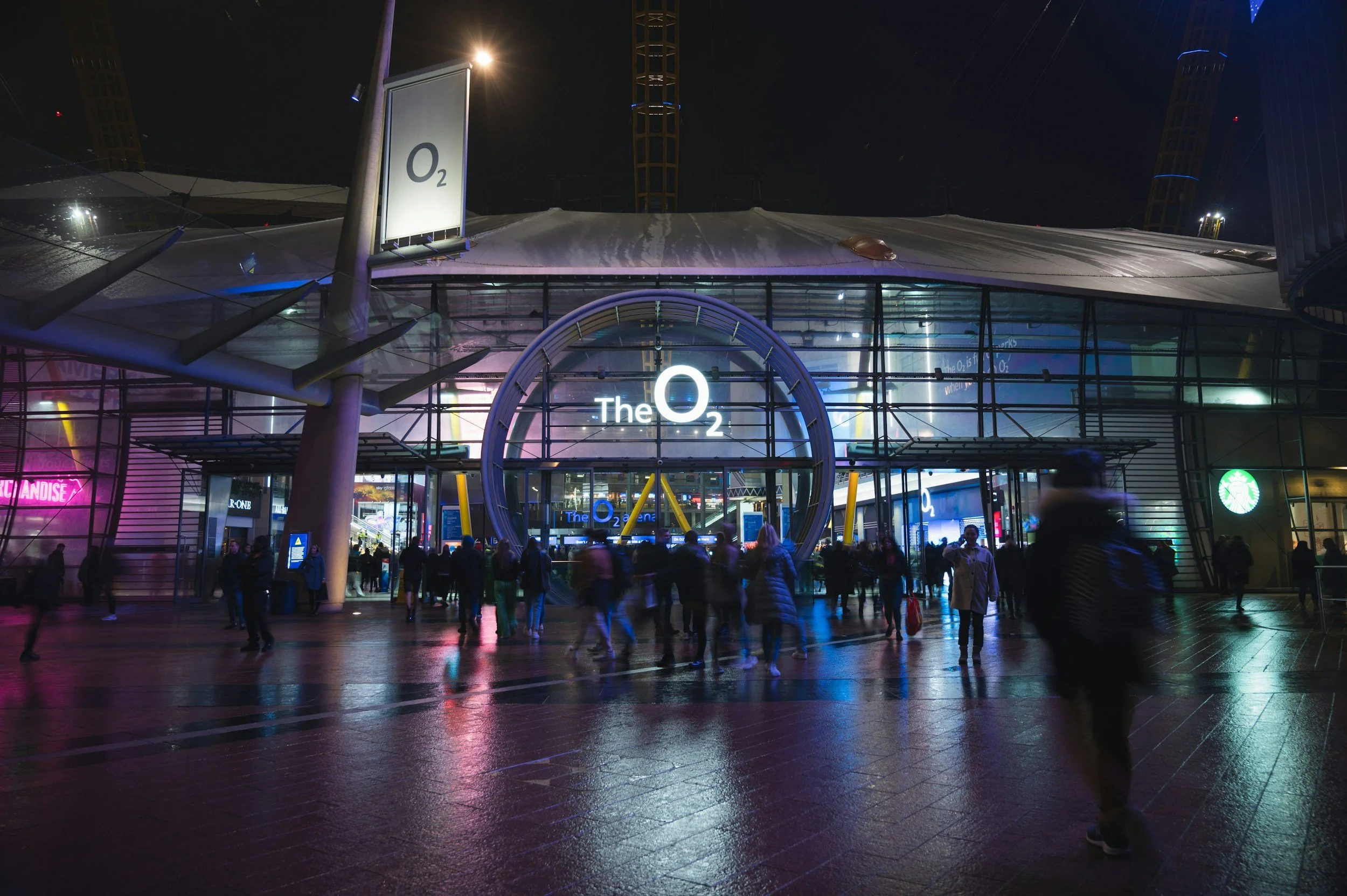 Nighttime view of the entrance to The O2, a large entertainment complex with a glass facade illuminated by colorful lights, crowded with people walking in and out.