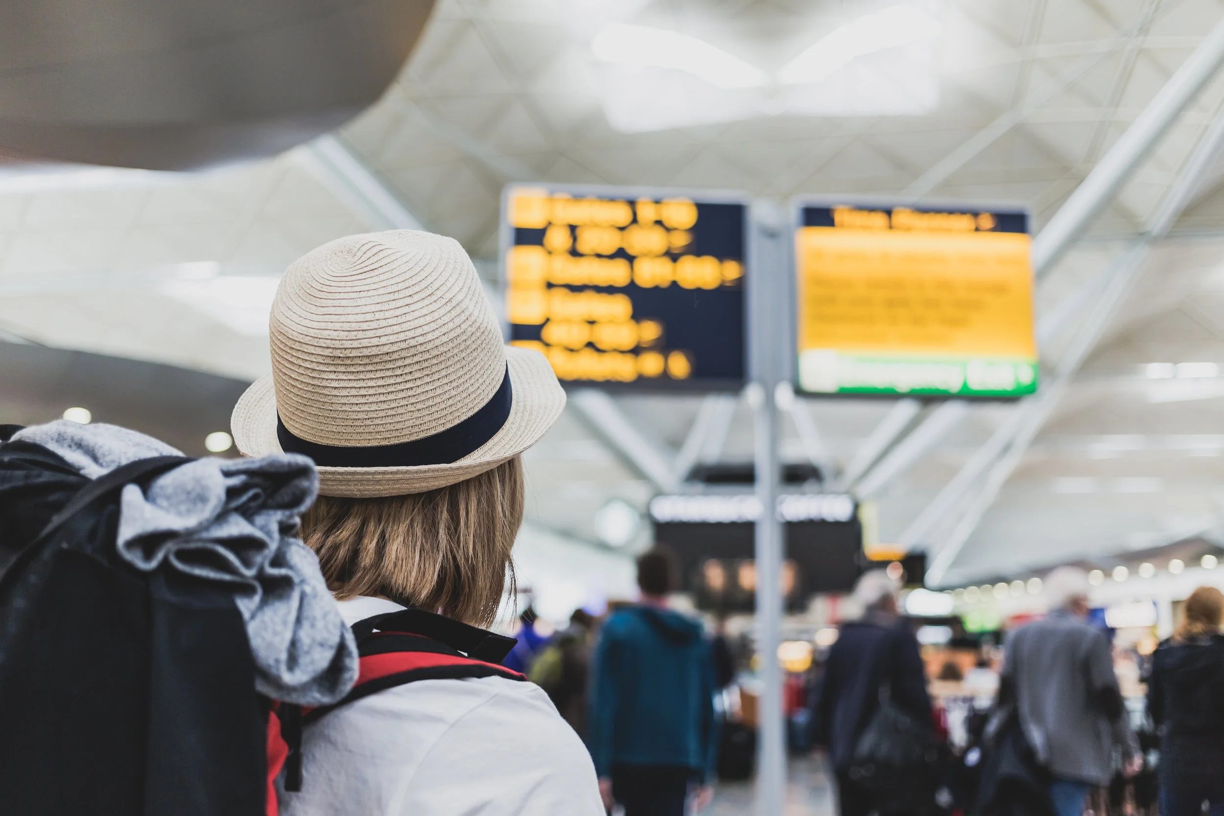 Taxis for travellers in West Malling, Kent, A person wearing a beige hat and a backpack at an airport with a crowded terminal and flight information screens in the background.