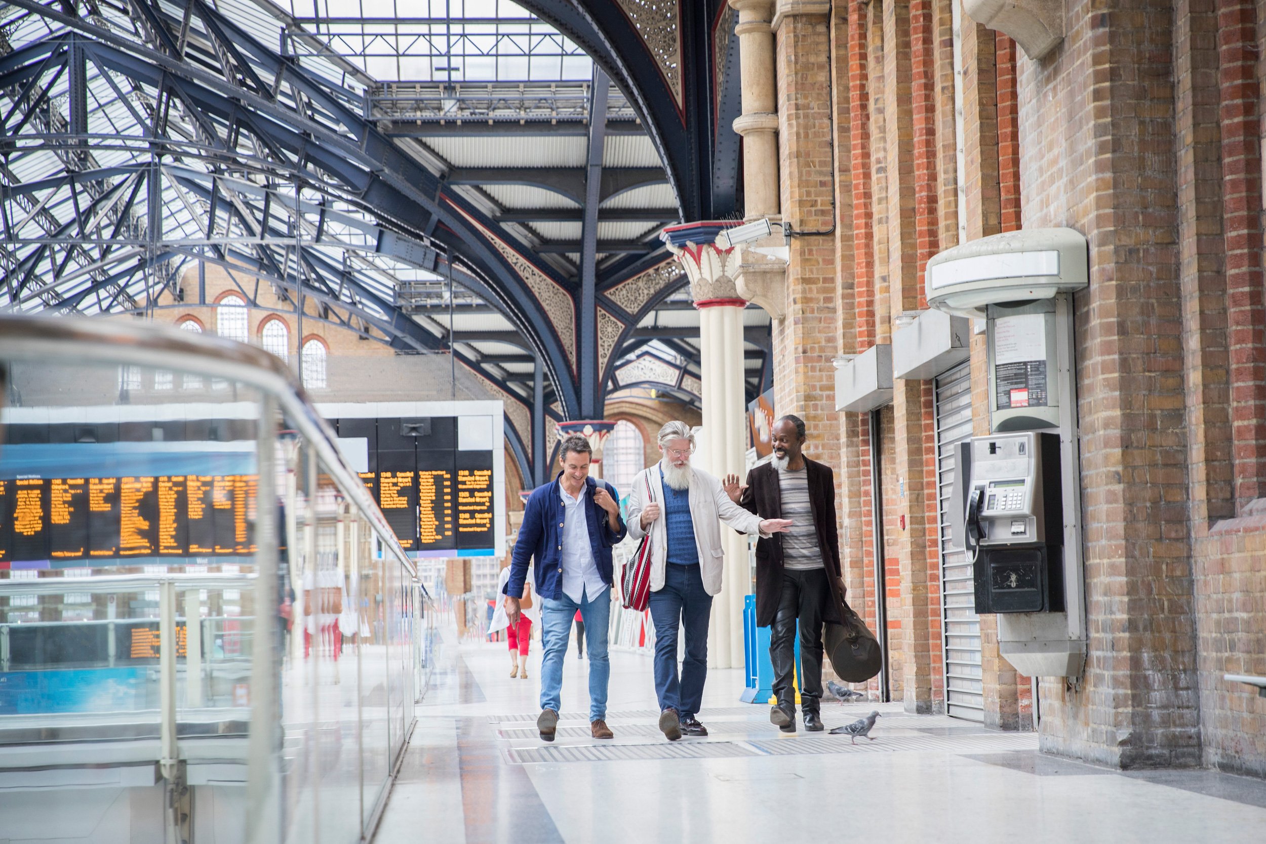 A group of four diverse people walking and talking inside a train station with brick walls, metal arches, and a digital departure board.