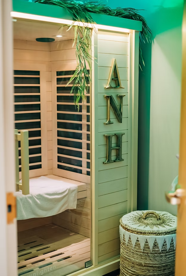 A sauna with wooden interior and black heater walls, decorated with green foliage, and a wicker laundry basket nearby.