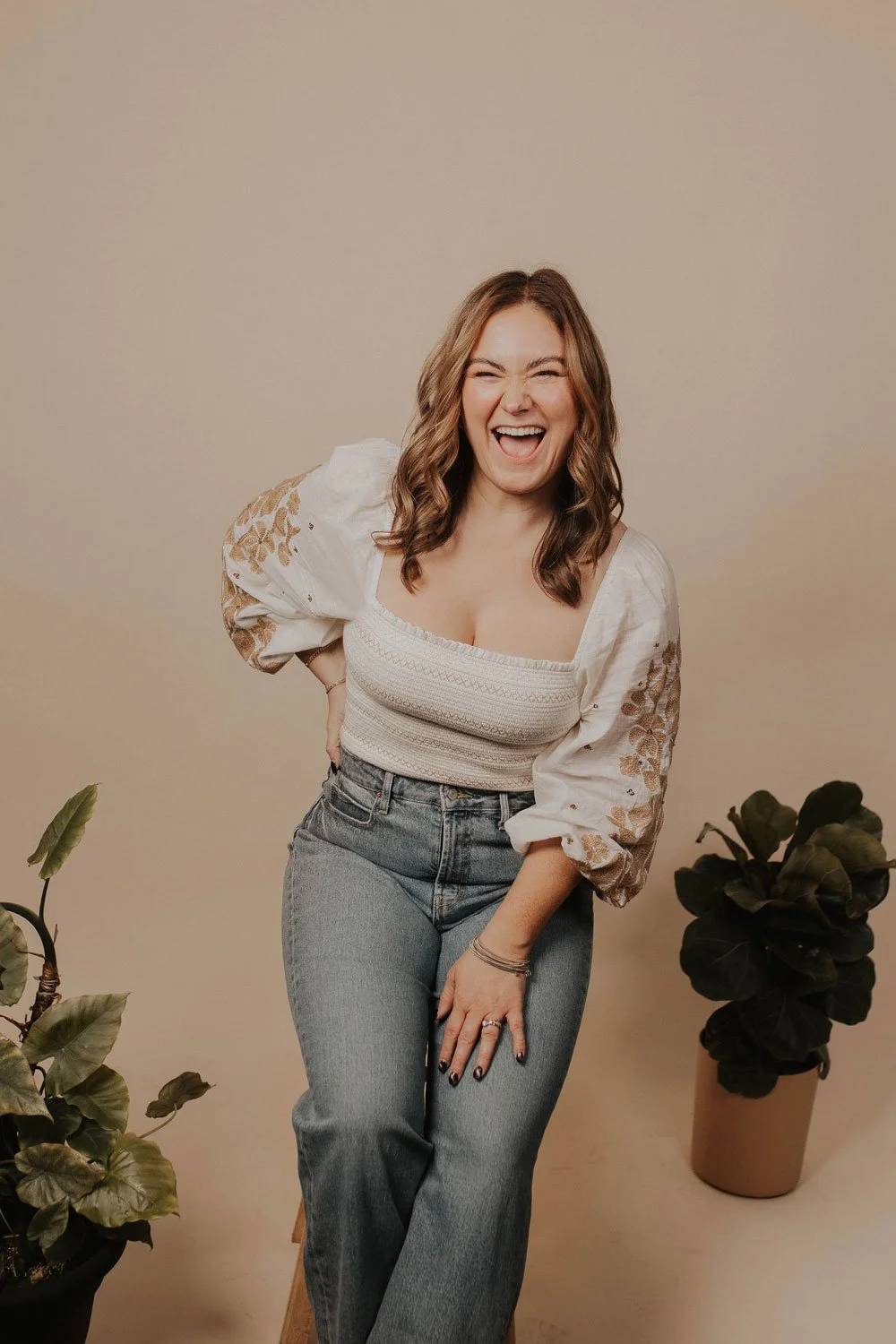 A woman with brown, wavy hair is smiling and laughing, posing with one hand on her hip in front of a plain beige background. She is wearing a white, embroidered blouse with puffed sleeves and high-waisted jeans. There are two potted plants on either side of her, one with large dark green leaves and the other with broad green and yellow leaves.