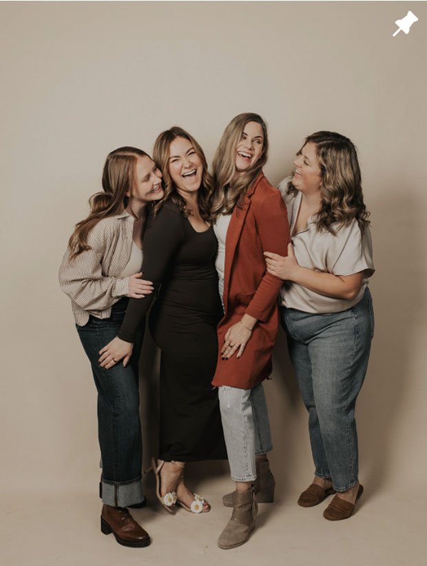 Four women laughing together against a plain background, dressed casually.