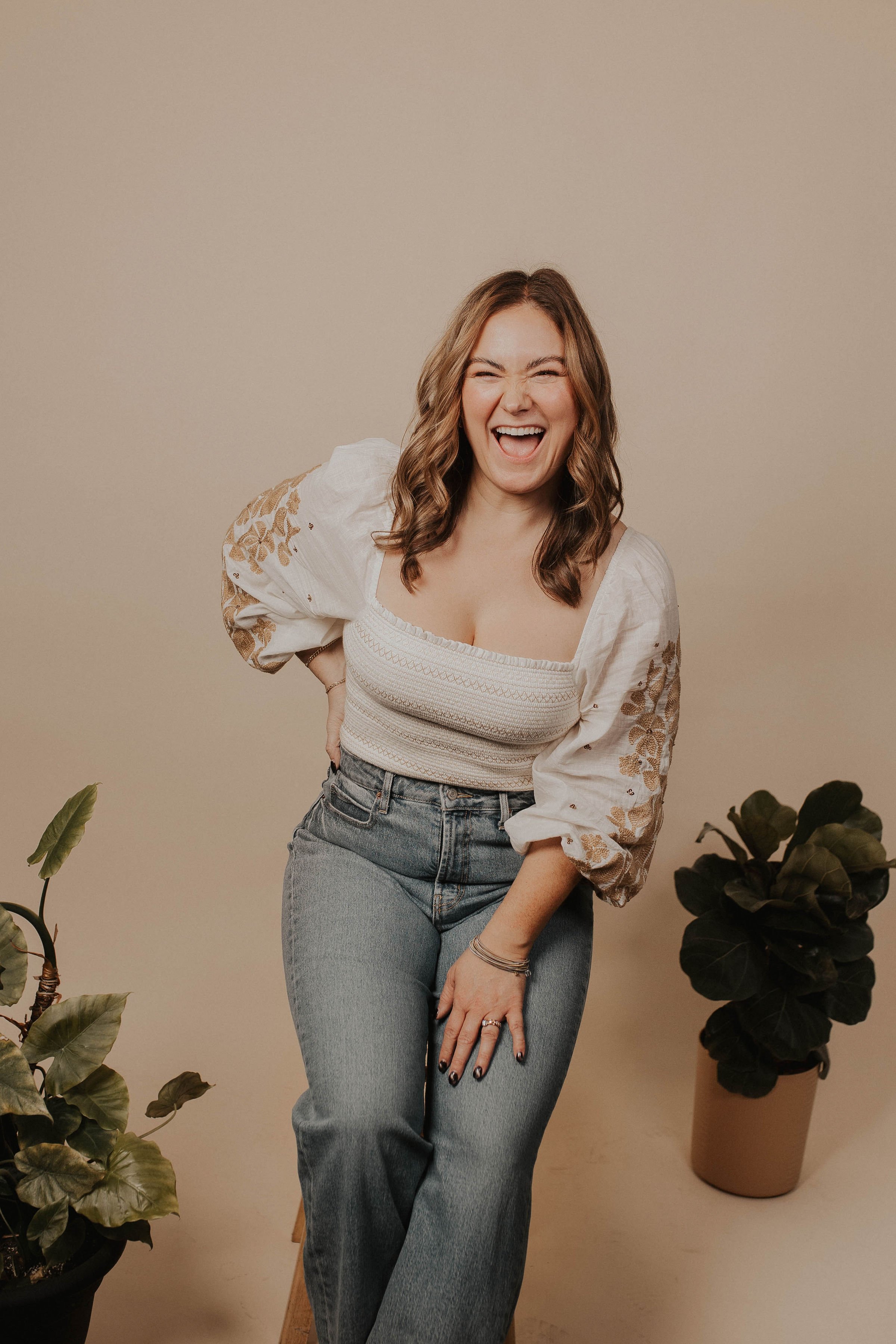 A woman with wavy hair laughing and posing with her right hand on her hip and left hand on her leg, wearing a cream blouse with embroidered sleeves and high-waisted jeans, standing against a beige background with potted plants on either side.