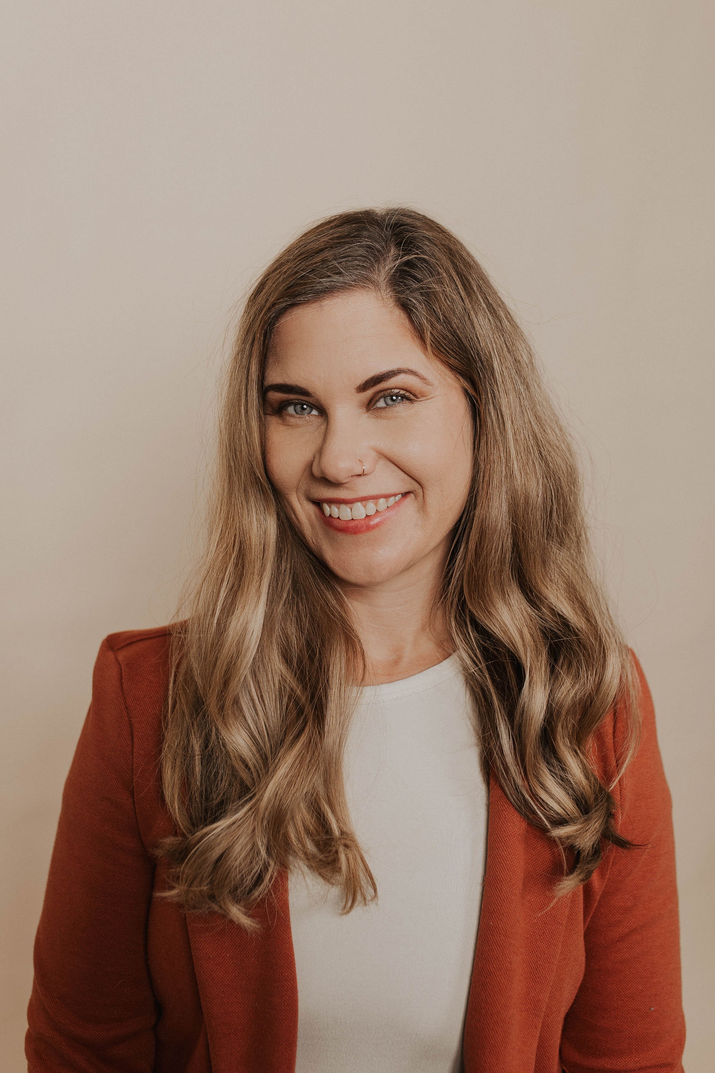 A woman with long, wavy light brown hair, smiling, wearing a white top and a rust-colored blazer, standing against a plain beige background.