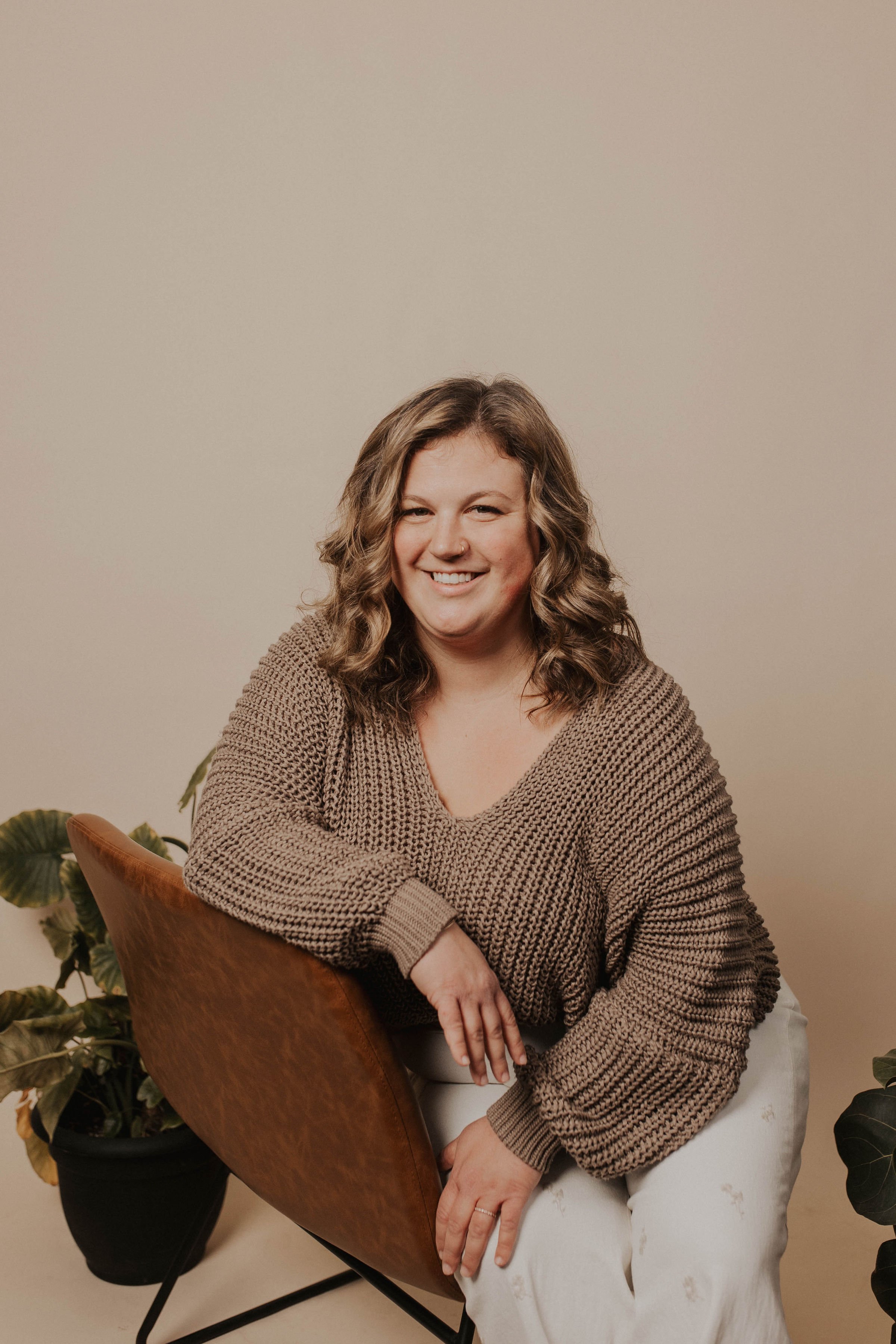 A smiling woman with curly hair wearing a beige sweater and white pants, sitting on a brown chair with plants nearby against a plain wall.