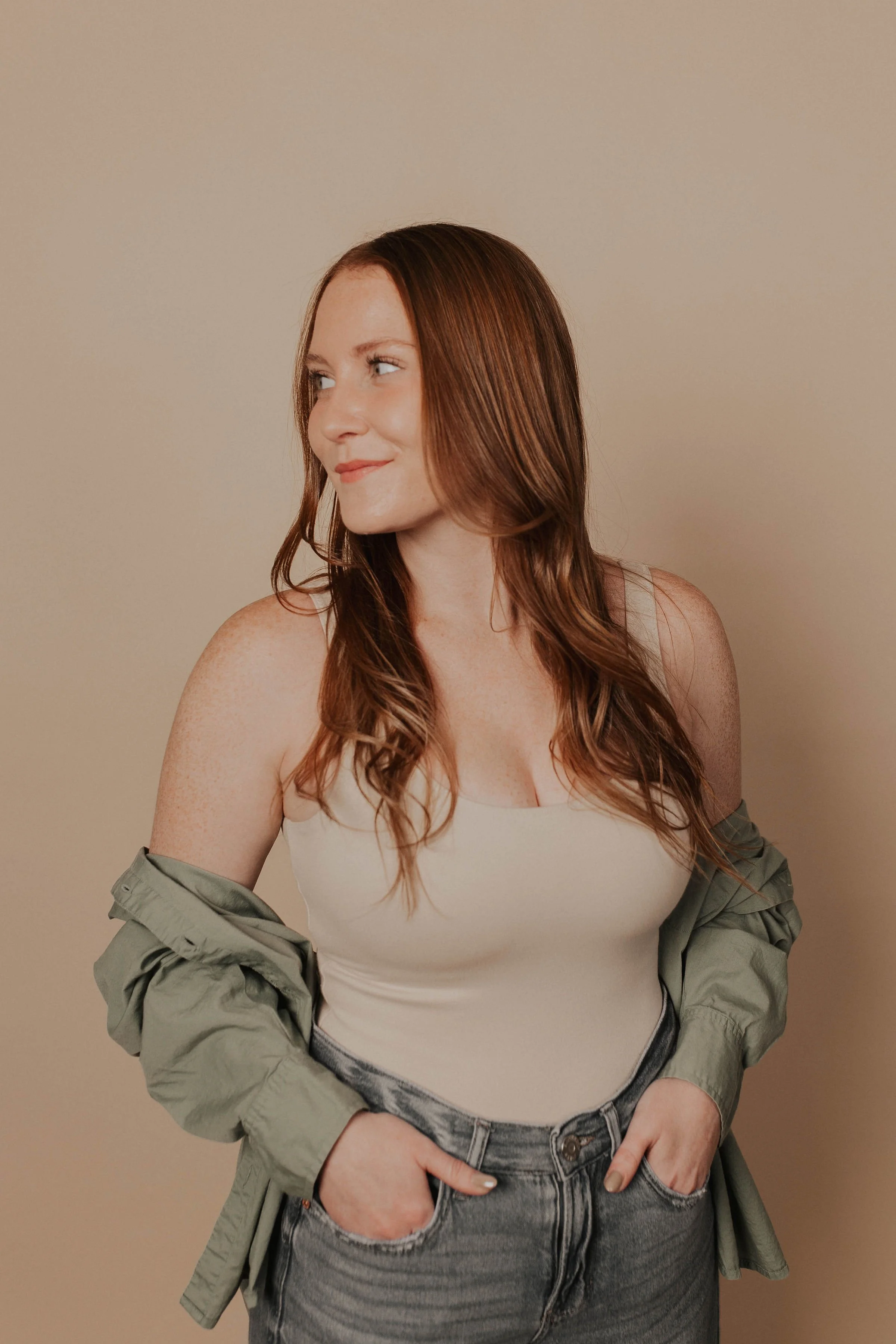A woman with long, wavy red hair wearing a cream tank top, gray jeans, and an olive green jacket draped over her shoulders, standing against a beige background.