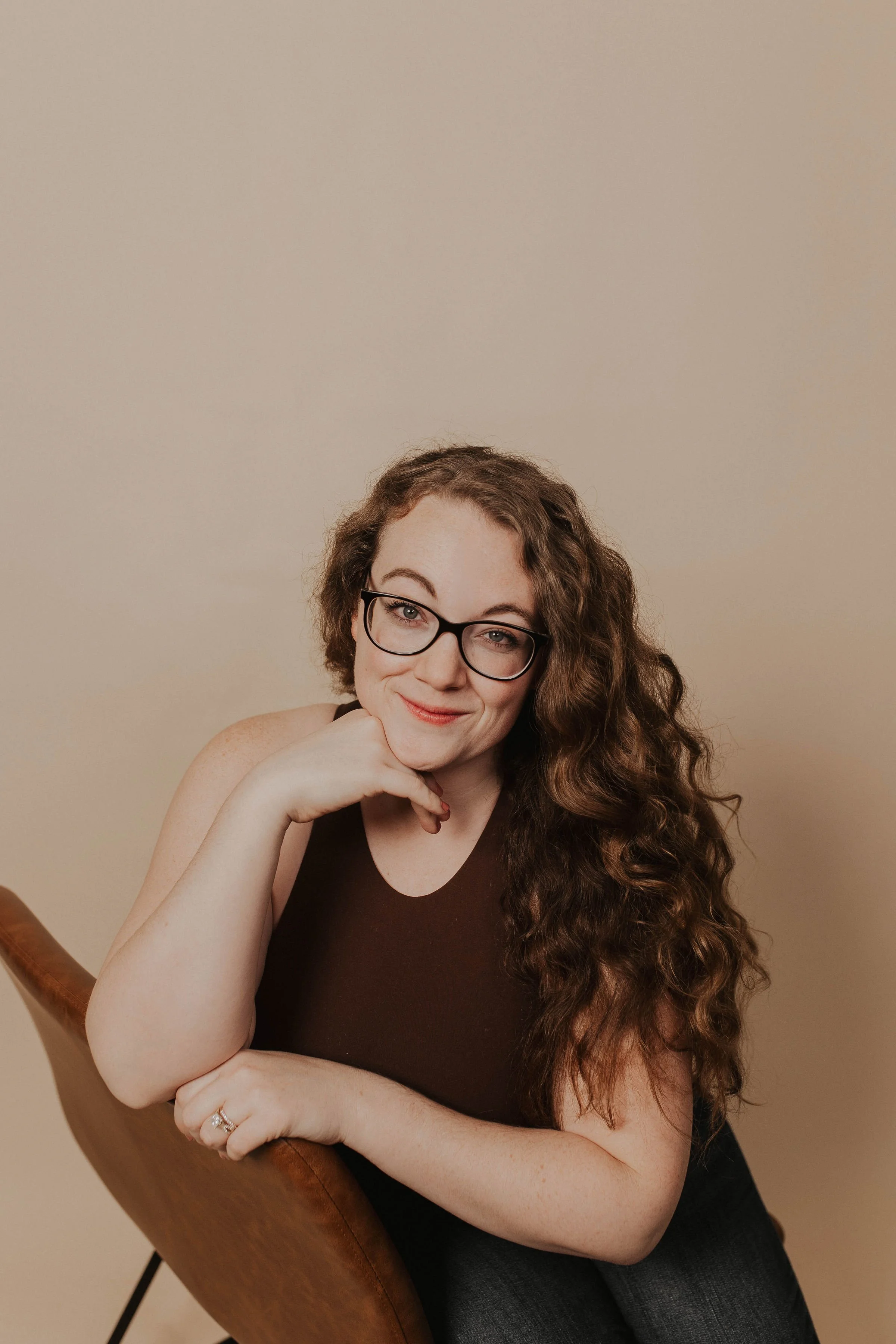 A woman with long, curly brown hair and glasses, smiling confidently, resting her chin on her hand, sitting on a brown chair against a beige background.