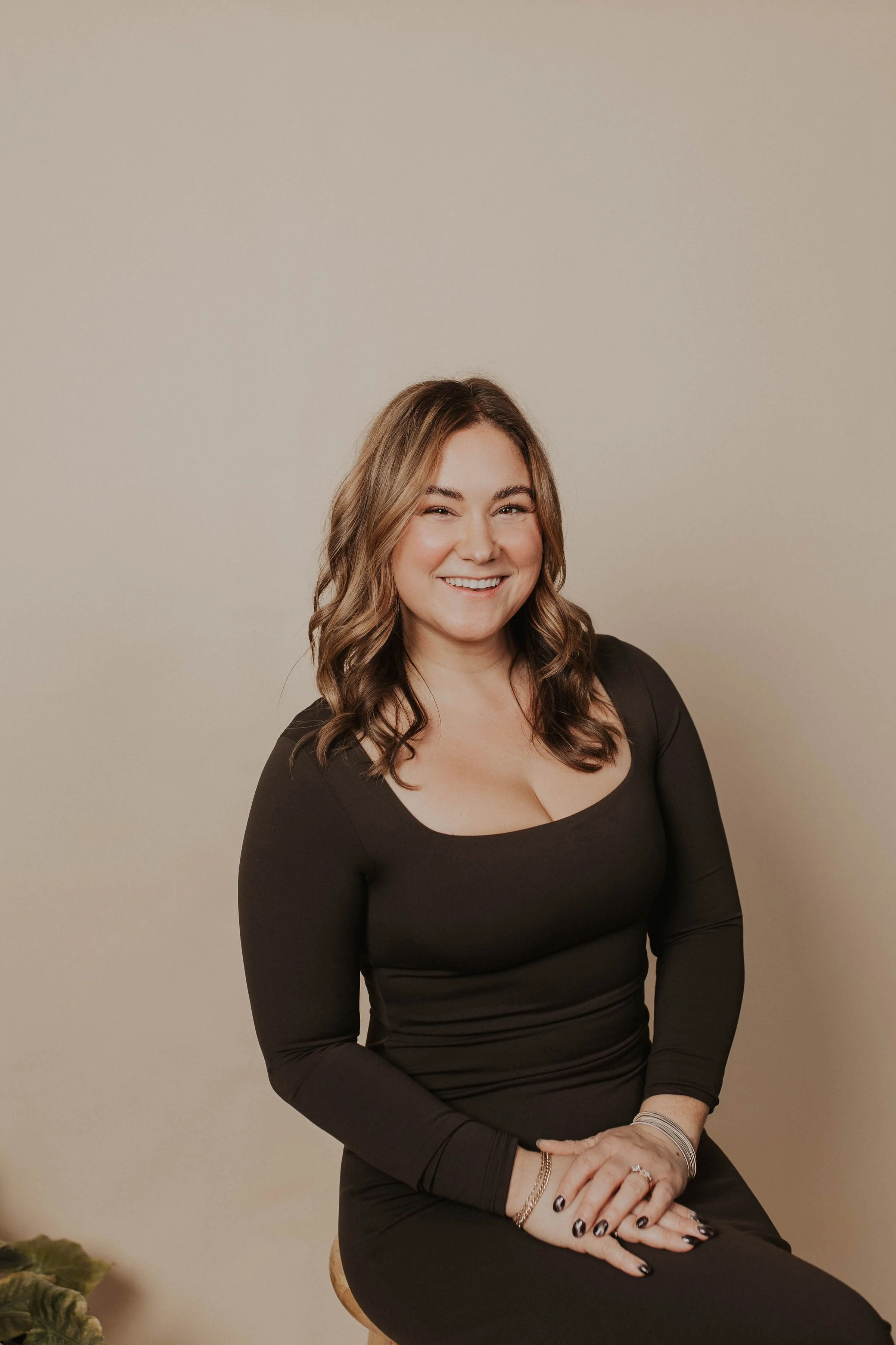 A woman with shoulder-length wavy brown hair, smiling and sitting on a stool against a plain beige wall, wearing a black long-sleeve top and black nails.