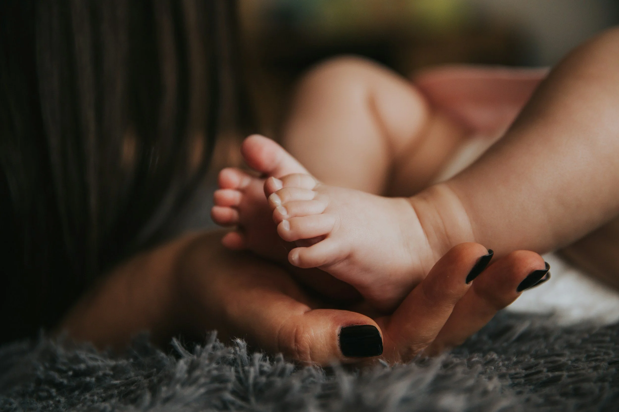 Close-up of an adult hand gently holding a baby's feet with toes curled, on a soft textured surface.