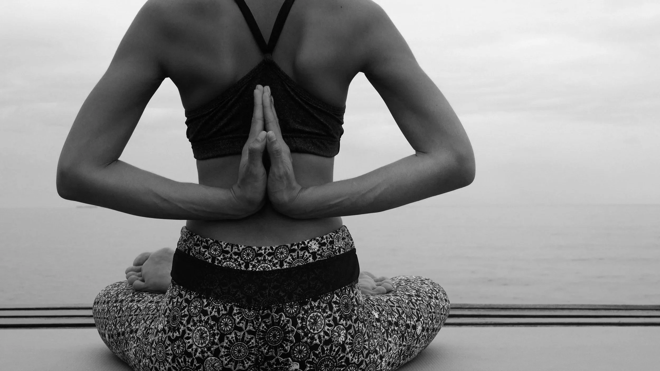 A person practicing yoga outdoors, sitting cross-legged on a mat, with hands in a prayer position behind their back, in front of an ocean or large body of water with a cloudy sky.