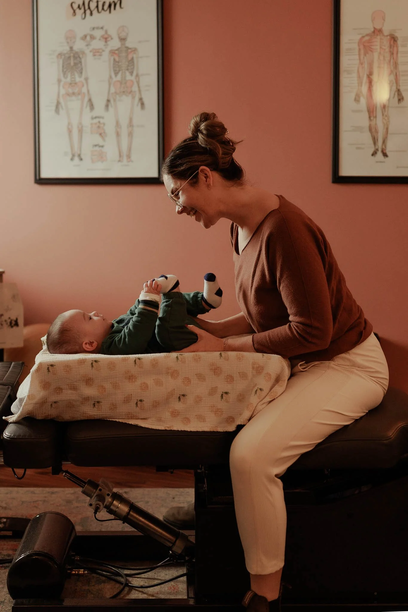 A woman smiling and leaning over a baby lying on a medical examination table, holding the baby's feet and legs. The baby is lying on a pillow with a floral pattern, wearing a green outfit, and looking up at the woman. The background shows anatomical posters on the wall.