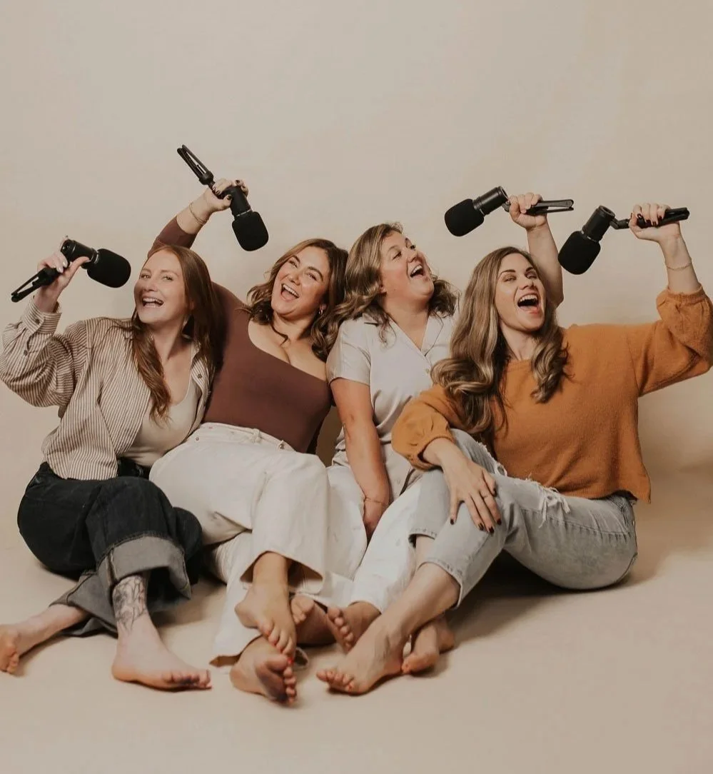 Four women sitting on the floor, holding microphones, smiling and laughing, against a plain background.