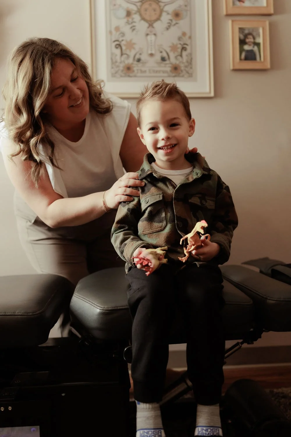 A woman and young boy smiling, sitting on a massage chair in a home, holding toy dinosaurs, with framed pictures and artwork on the wall behind them.