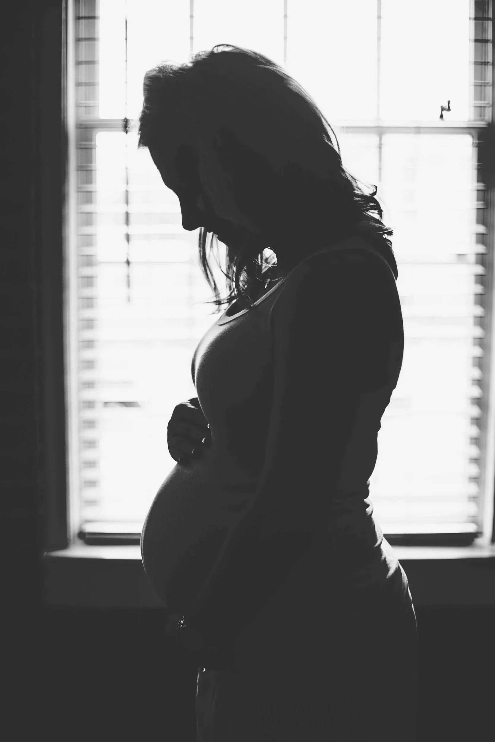 Silhouette of a pregnant woman standing in front of a bright window, looking down with her hand on her pregnant belly.