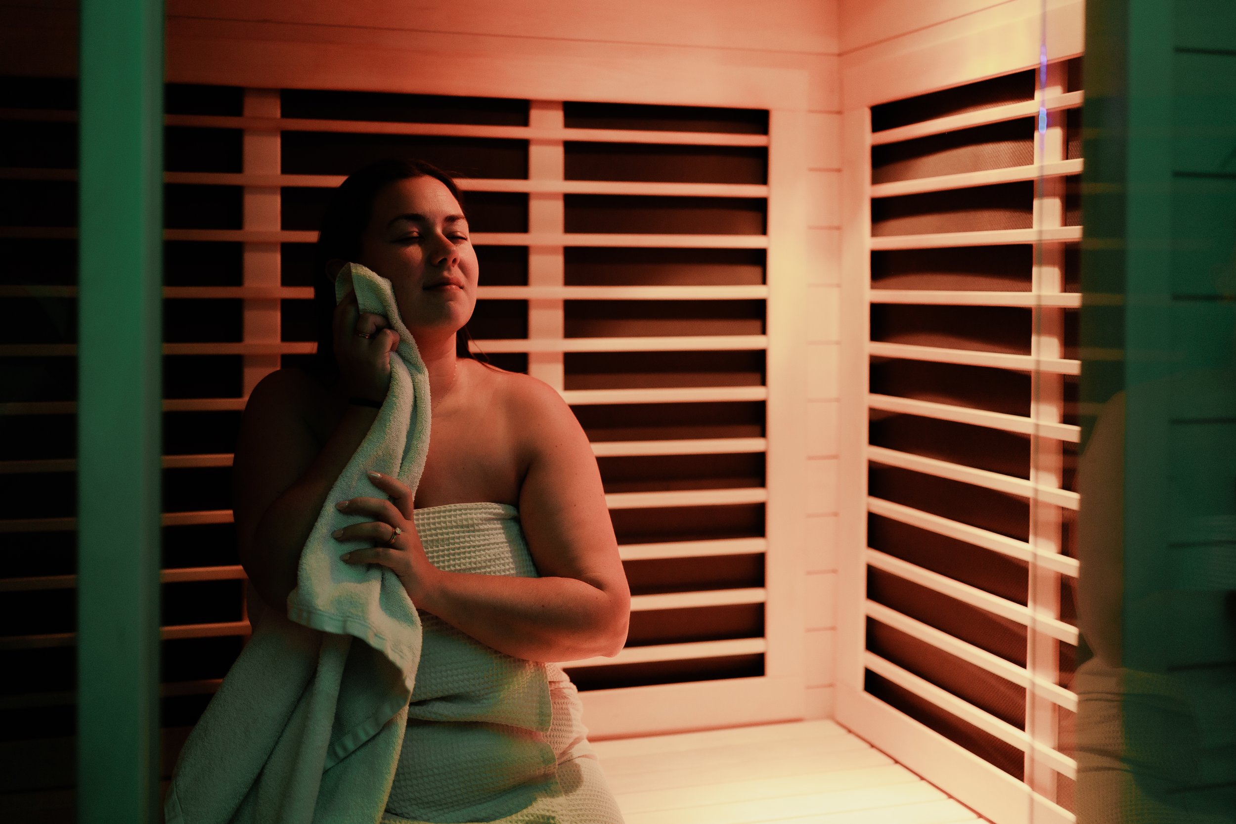 A woman with eyes closed, holding a towel to her face, sitting inside a wooden sauna room.