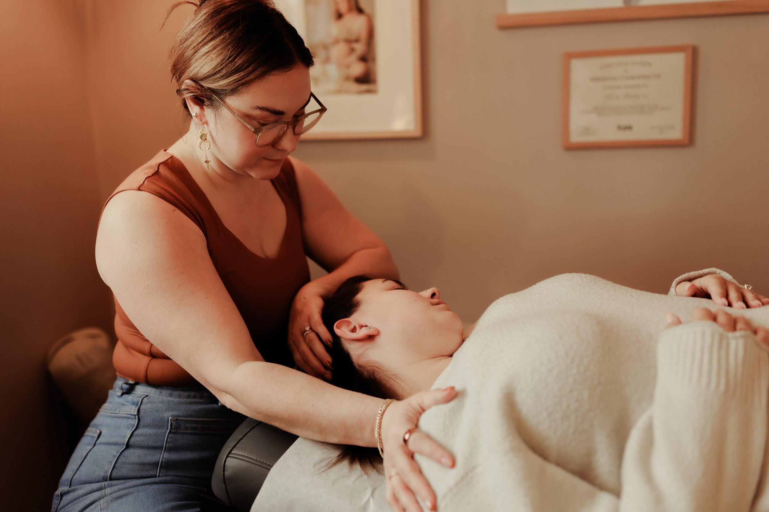 A woman with glasses and earrings gently supports and comforts another woman lying on a massage table. The woman on the table has her eyes closed and is wrapped in a white blanket. The room has warm lighting and framed certificates or documents on the wall.