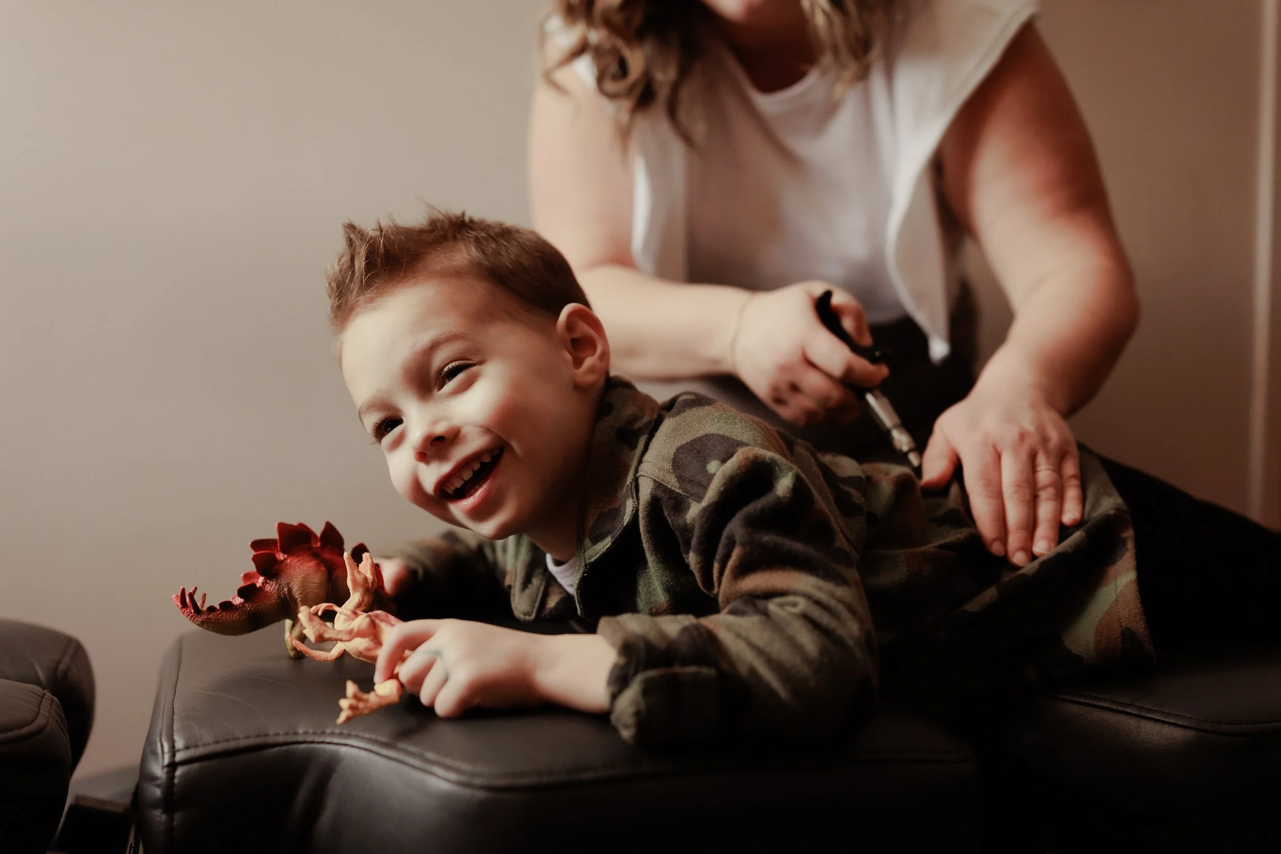 A young boy in camouflage clothing laughing as he lies on a black leather ottoman, holding a toy dinosaur, while a woman, possibly his mother, prepares to give him a shot.