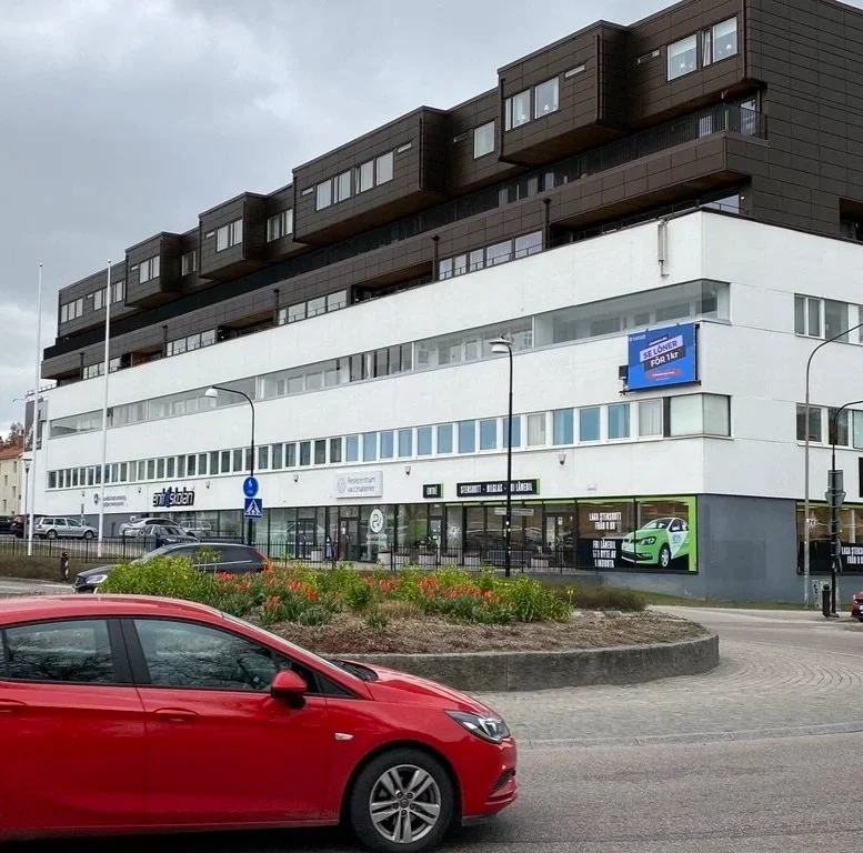 Moderna, flerfärgade kontors- och bostadsbyggnader med bilar parkerade framför, en blomsterbädd, och skyltar för butiker.