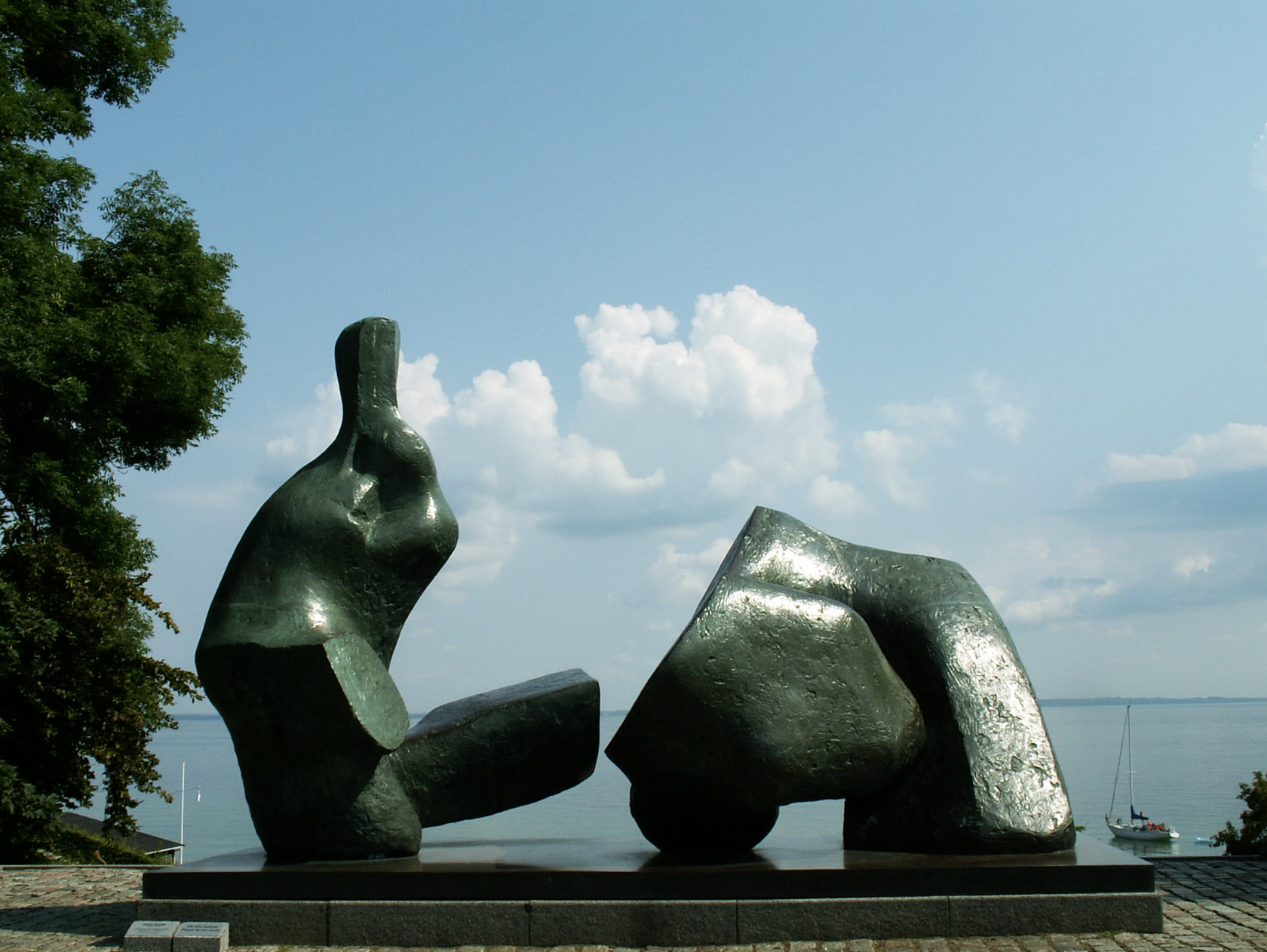 Large abstract metallic sculpture with three parts, set against a view of water with a sailboat, trees, and a cloudy sky in the background.