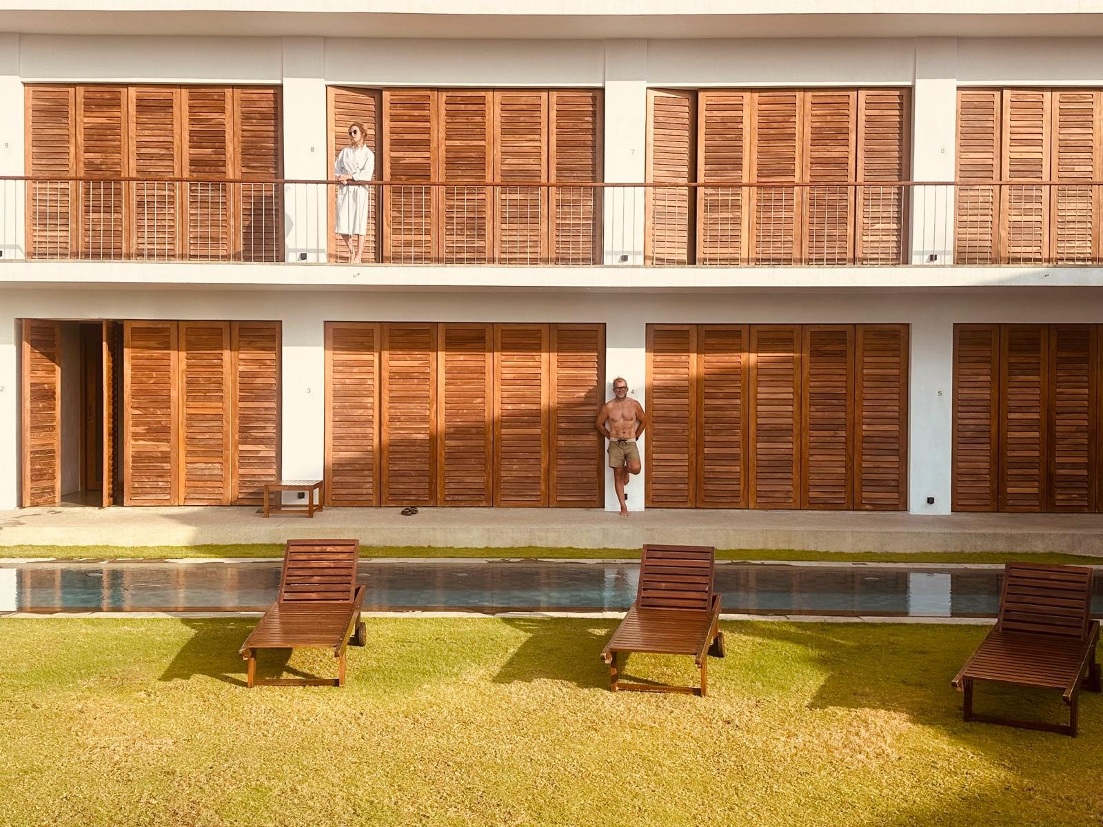 A woman in a white dress and sunglasses standing on a balcony, a shirtless man in shorts leaning against a wall, three wooden lounge chairs on a grass lawn next to a pool, and a modern building with wooden shutters.