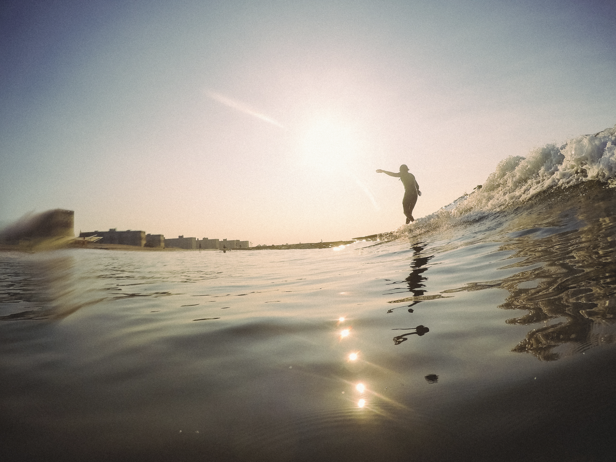 A surfing on at sunset with the sun behind them over New York and Rockaway Beach. The water is calm with gentle ripples, and the coastline with buildings is visible in the distance.