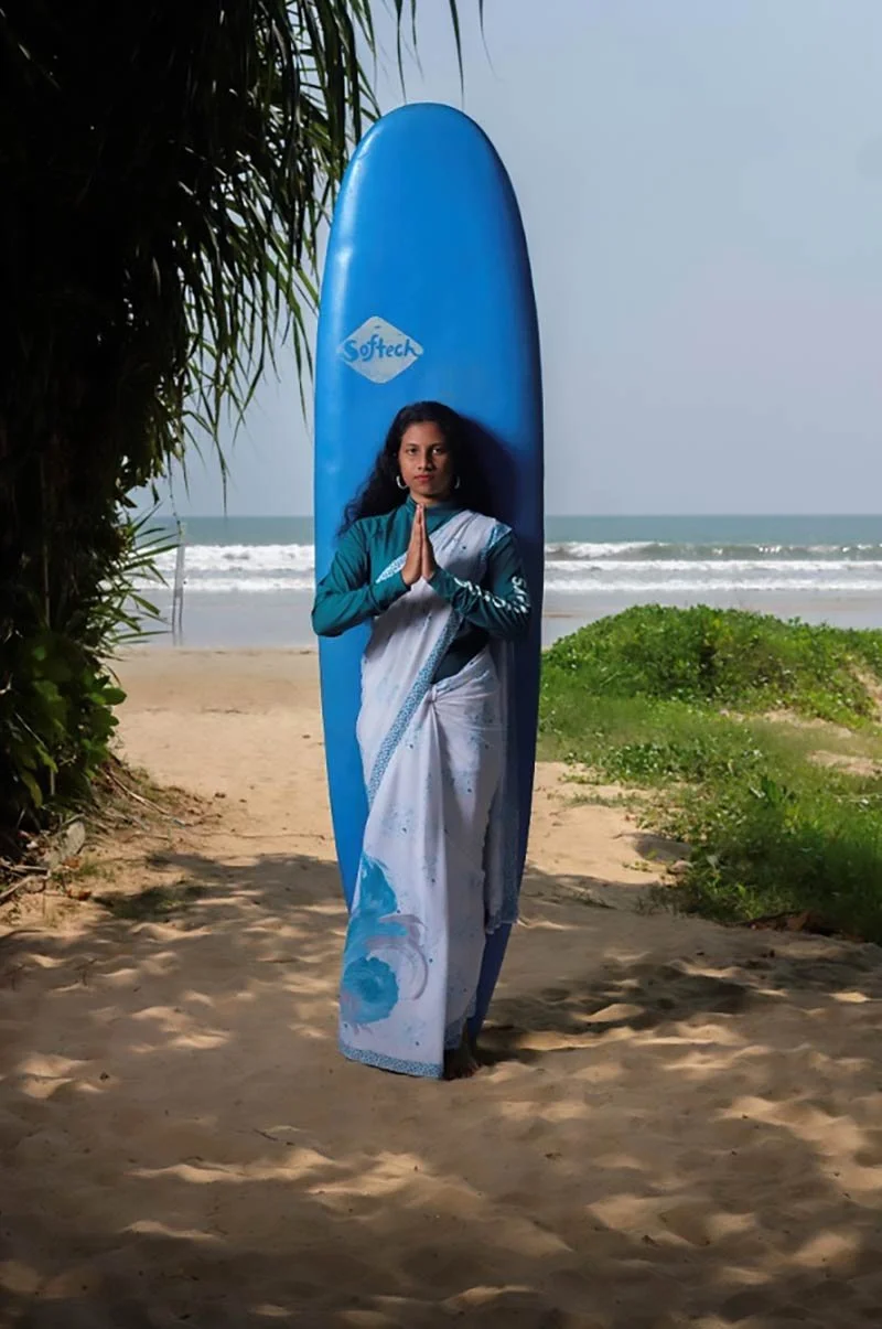 A woman in a white saree with blue accents standing on a sandy beach, holding her hands together in a prayer position in front of an upright blue surfboard with a 'Softech' logo, near the shoreline with ocean waves in the background.