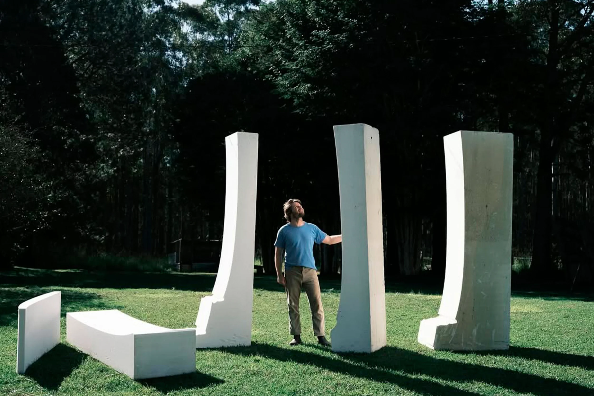 Ellis standing in a grassy park area surrounded by large abstract white planks, with trees in the background.