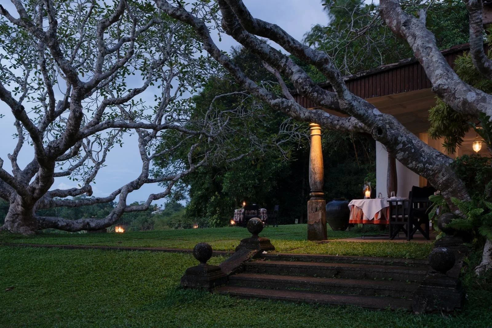A cozy outdoor dining area on a porch at dusk, illuminated by lanterns, with a large tree and a grassy yard in the background.