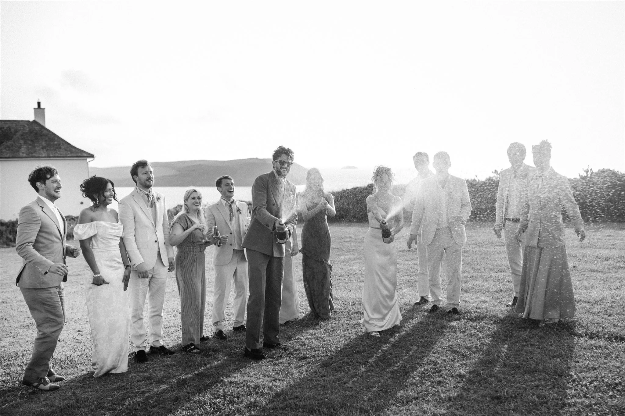 A group of people dressed in formal attire celebrating outdoors, holding bottles and spraying champagne, with a house and landscape in the background.
