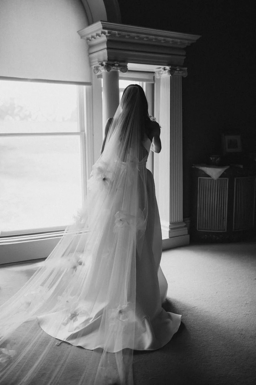 A bride standing indoors near a large window, dressed in a wedding gown with a long veil, looking outside.
