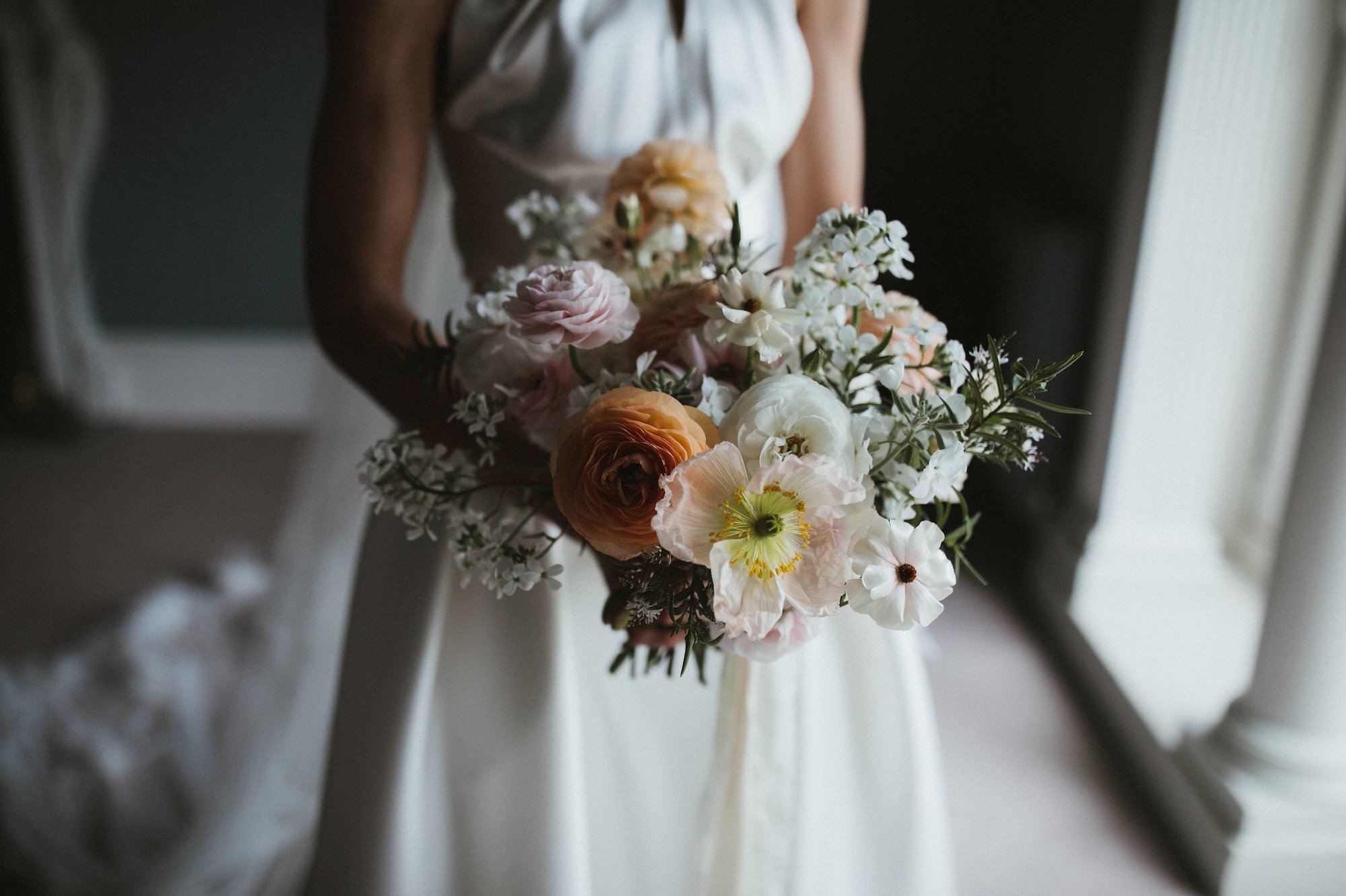 A woman in a white dress holding a bouquet of assorted pastel-colored flowers, including pink, white, peach, and cream blooms, near a window with light coming through.