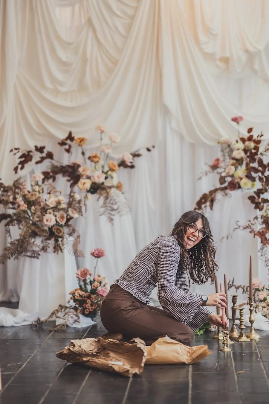 your wedding planner arranging candles on a floor in front of floral arrangements and draped white fabric at an event or wedding setup.