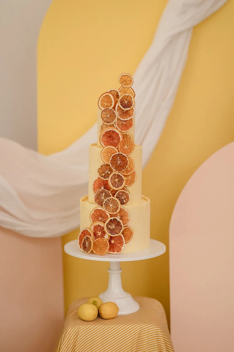 Three-tiered cake decorated with dried citrus slices, placed on a white cake stand. The background features pastel yellow and pink colors, with a small tablecloth and three whole apples in front of the cake.