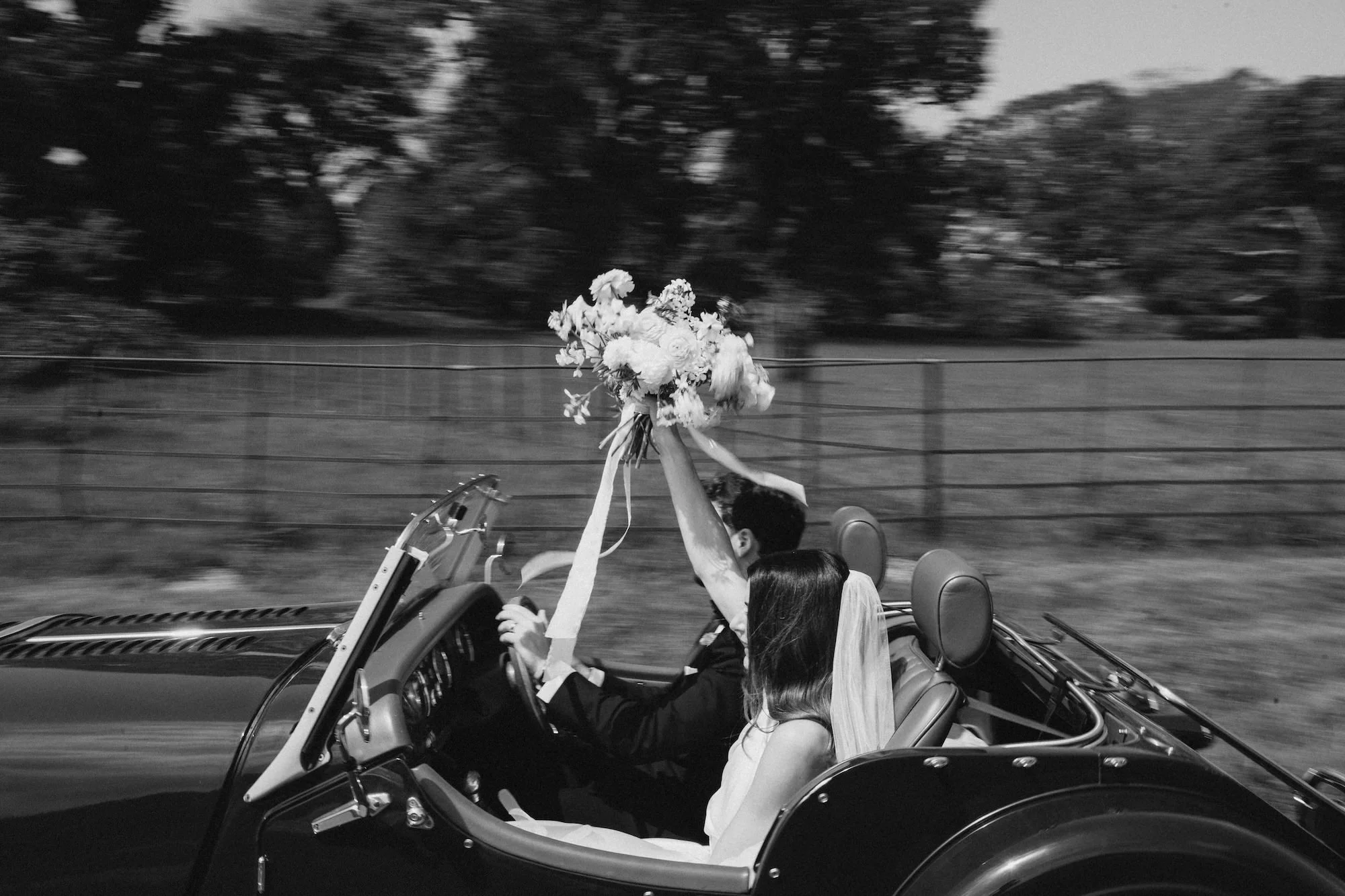 Black and white photo of a couple in a vintage convertible car, with the bride holding a bouquet of flowers up in the air as they drive down a road.