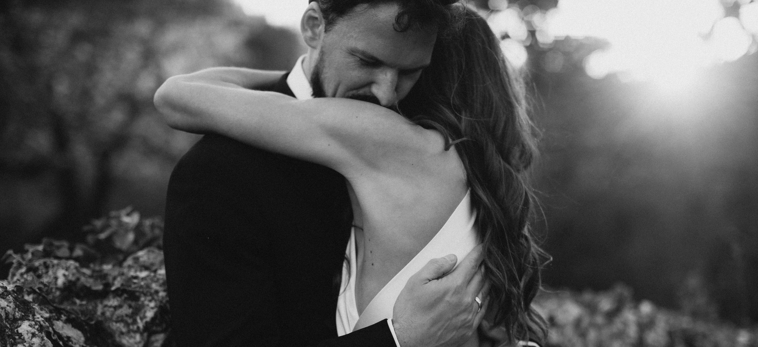 Groom embraces a woman in a white dress outdoors, with trees and sunlight in the background.