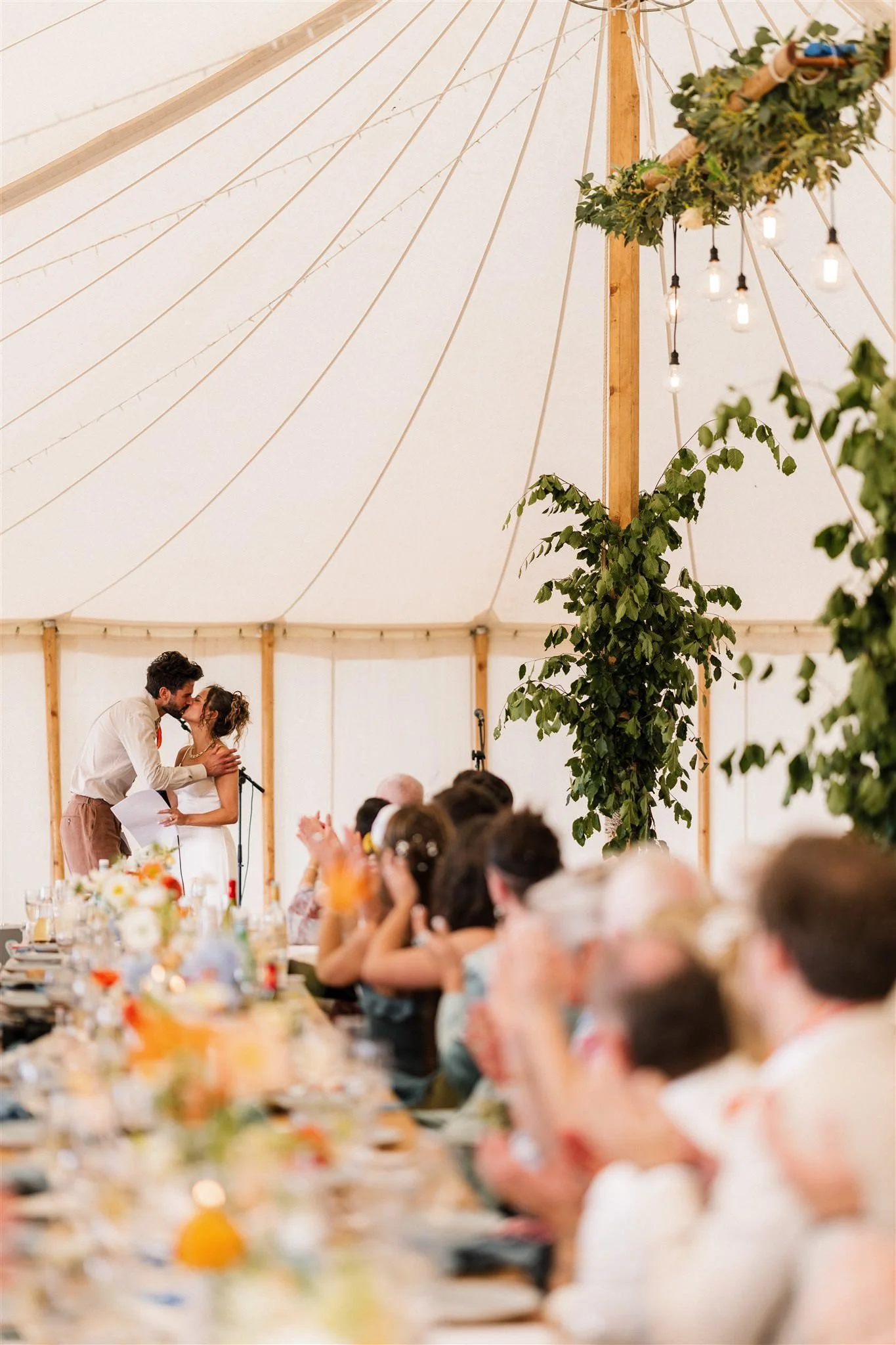 A wedding reception inside a large tent with a couple kissing at the head table, surrounded by family and friends, decorated with green foliage and string lights.