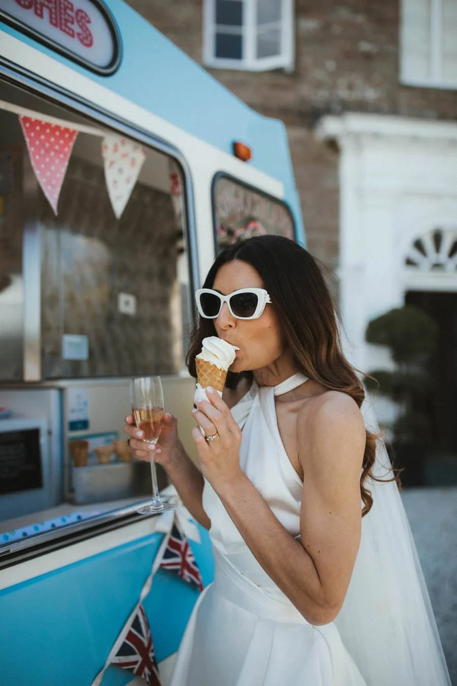 A woman in a white dress and large sunglasses enjoys ice cream and champagne in front of a vintage ice cream truck.