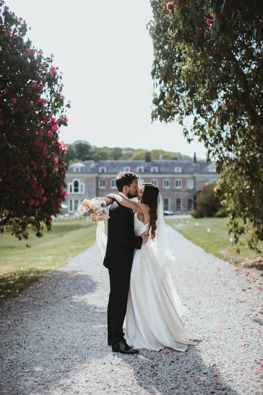 A newlywed couple sharing a kiss outdoors on a gravel path surrounded by trees and a large historic building in the background, with the bride holding a bouquet of flowers.